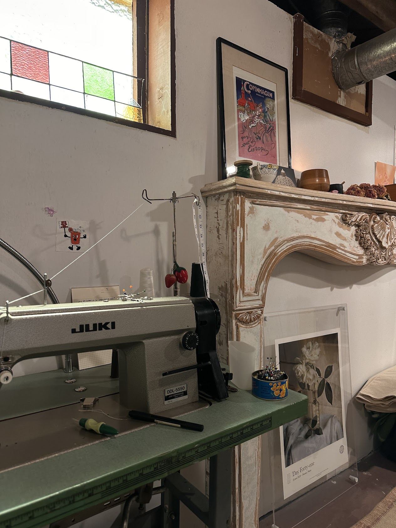 An industrial sewing machine on a green table beside a carved mantel and framed prints in the basement studio, with the original metal duct visible near the ceiling